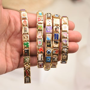Könnte beinhalten: Goldfarbene Charm-Armbänder mit verschiedenen bunten Emaille-Anhängern. Die Anhänger zeigen Symbole, Wörter und Designs. Die Armbänder werden in einer Hand gehalten und zeigen die verschiedenen Stile.