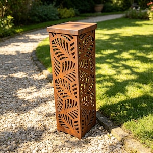 Può includere: Un pilastro da giardino rettangolare in metallo dall'aspetto usurato con una parte superiore quadrata. Il pilastro presenta un design traforato a forma di foglie. Il colore è marrone ruggine e si trova su un sentiero di ghiaia in un giardino.