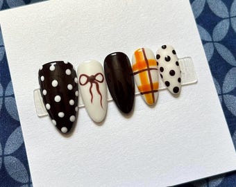 May include: A set of five artificial nails with various designs. The nails feature a dark brown base with white polka dots, a white base with a brown bow, a solid dark brown nail, a plaid pattern in orange and white, and a white base with black polka dots.