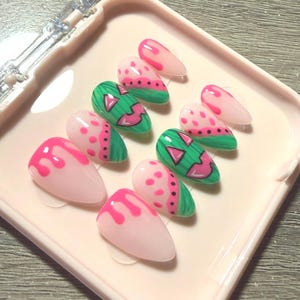 May include: A set of ten press-on nails featuring a watermelon-inspired design. The nails display a mix of translucent pink bases with bright pink drip patterns, green sections with black seeds, and green segments with pink triangular watermelon slices.