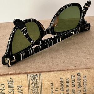 May include: Vintage black sunglasses with a white marbled pattern and green lenses. The sunglasses are resting on a vintage book titled "Flying Yorkshire-Man" by Eric Knight. The book has a tan cover with black text.