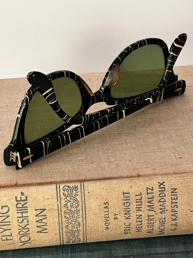 May include: Vintage black sunglasses with a white marbled pattern and green lenses. The sunglasses are resting on a vintage book titled "Flying Yorkshire-Man" by Eric Knight. The book has a tan cover with black text.