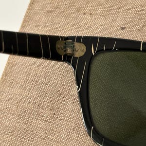 May include: Close-up of black sunglasses with white stripe accents. The sunglasses have a rectangular shape with green lenses. The hinges are a brass color. The sunglasses are resting on a tan fabric surface.