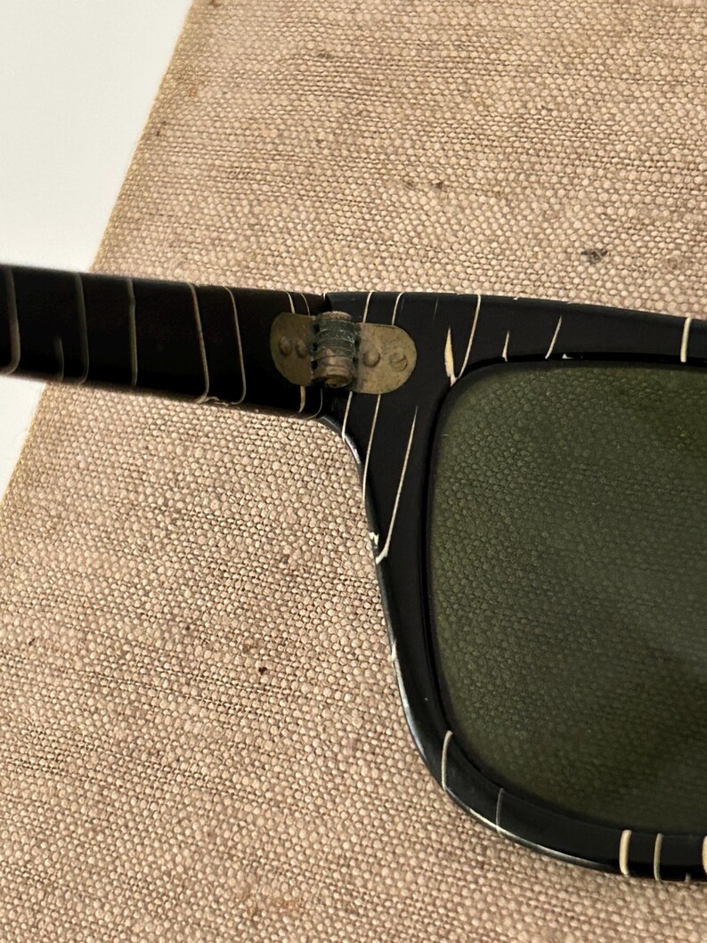 May include: Close-up of black sunglasses with white stripe accents. The sunglasses have a rectangular shape with green lenses. The hinges are a brass color. The sunglasses are resting on a tan fabric surface.