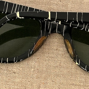 May include: Close-up of black sunglasses with a white striped pattern. The sunglasses have dark green lenses and a rectangular frame. The arms of the sunglasses are also black with the same white striped pattern.