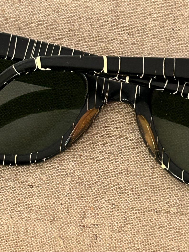 May include: Close-up of black sunglasses with a white striped pattern. The sunglasses have dark green lenses and a rectangular frame. The arms of the sunglasses are also black with the same white striped pattern.