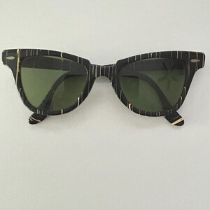 May include: Black sunglasses with a striped pattern and green lenses. The frames have a retro design with a keyhole bridge. The sunglasses are set against a white background.