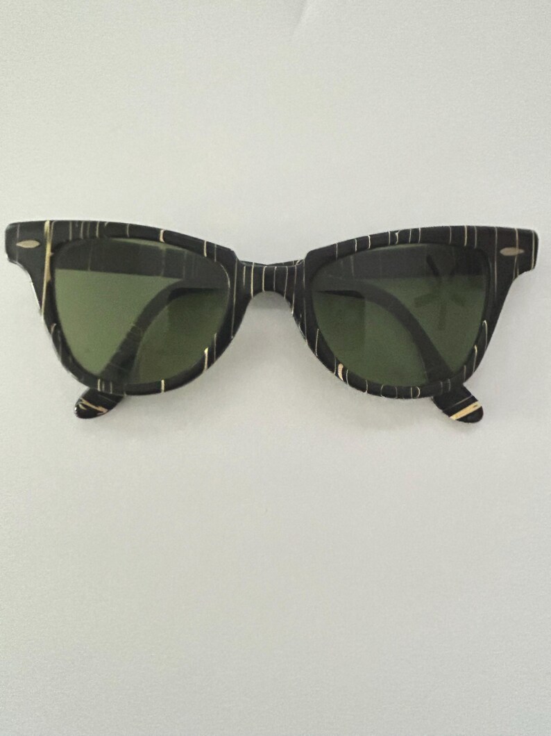 May include: Black sunglasses with a striped pattern and green lenses. The frames have a retro design with a keyhole bridge. The sunglasses are set against a white background.