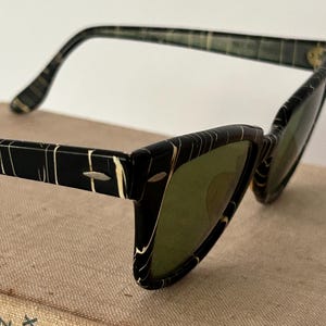 May include: Vintage cat-eye sunglasses with a black and white marbled pattern frame and green lenses. The sunglasses have a retro design, with a sleek, angular shape. The arms of the sunglasses are also black and white.