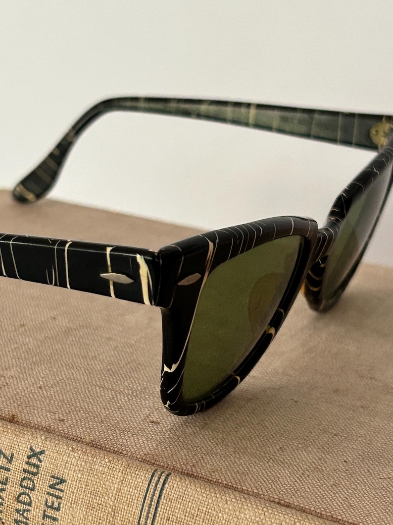 May include: Vintage cat-eye sunglasses with a black and white marbled pattern frame and green lenses. The sunglasses have a retro design, with a sleek, angular shape. The arms of the sunglasses are also black and white.