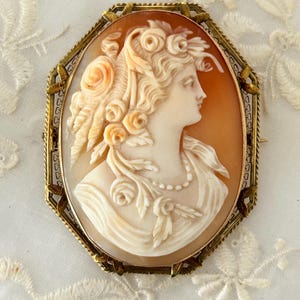 May include: An antique cameo brooch featuring a carved profile of a woman with floral accents in her hair and around her neck. The cameo is set in an octagonal gold-toned frame, showcasing intricate detailing and a classic aesthetic.