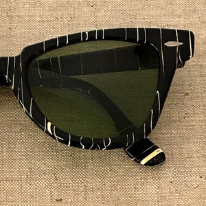 May include: Black sunglasses with a white striped pattern on the frames. The lenses are a dark green color. The sunglasses have a classic, retro design.