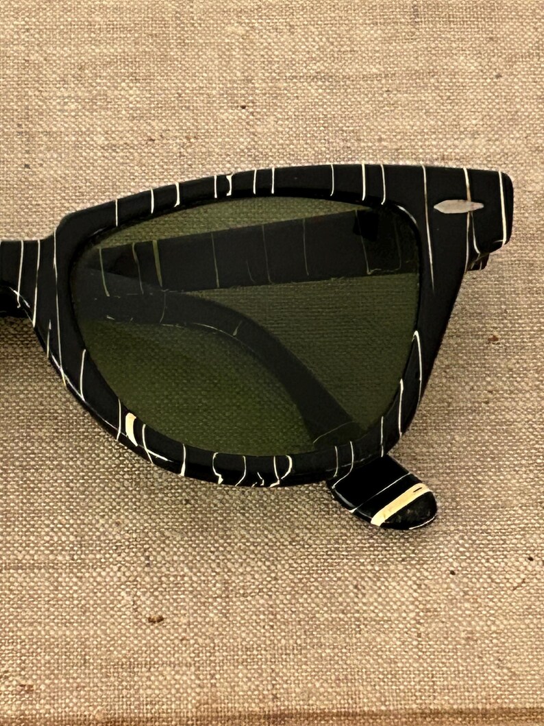 May include: Black sunglasses with a white striped pattern on the frames. The lenses are a dark green color. The sunglasses have a classic, retro design.