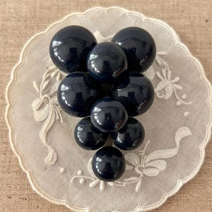 May include: A cluster of dark navy blue, glossy, spherical beads arranged in a grape-like shape. The beads are resting on a white, scalloped-edged doily with embroidered floral patterns. The background is a tan fabric.
