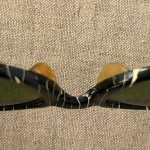 May include: Close-up of sunglasses with a black frame and a white and gold pattern. The lenses are green. The nose pads are a light brown color. The sunglasses are set against a neutral background.