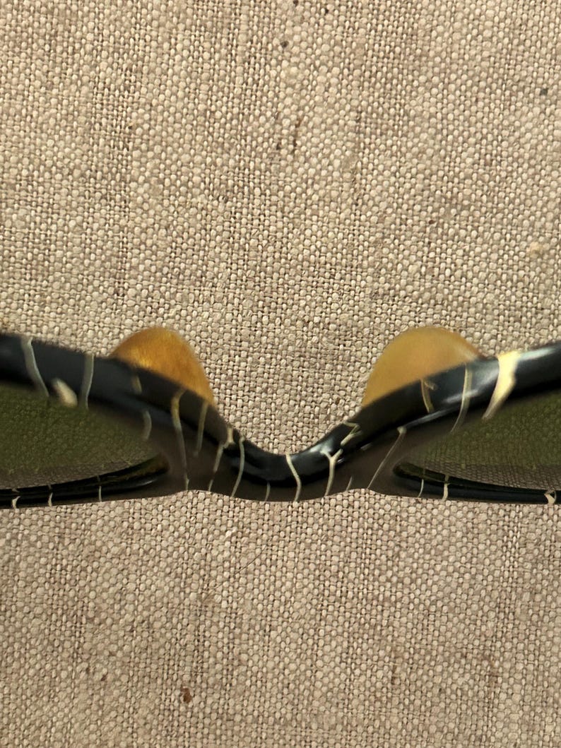 May include: Close-up of sunglasses with a black frame and a white and gold pattern. The lenses are green. The nose pads are a light brown color. The sunglasses are set against a neutral background.
