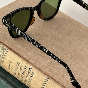 May include: Black cat-eye sunglasses with green lenses. The frames feature a unique pattern of white lines on a black background. The sunglasses are resting on a vintage book with the title "Novellas" and author names.