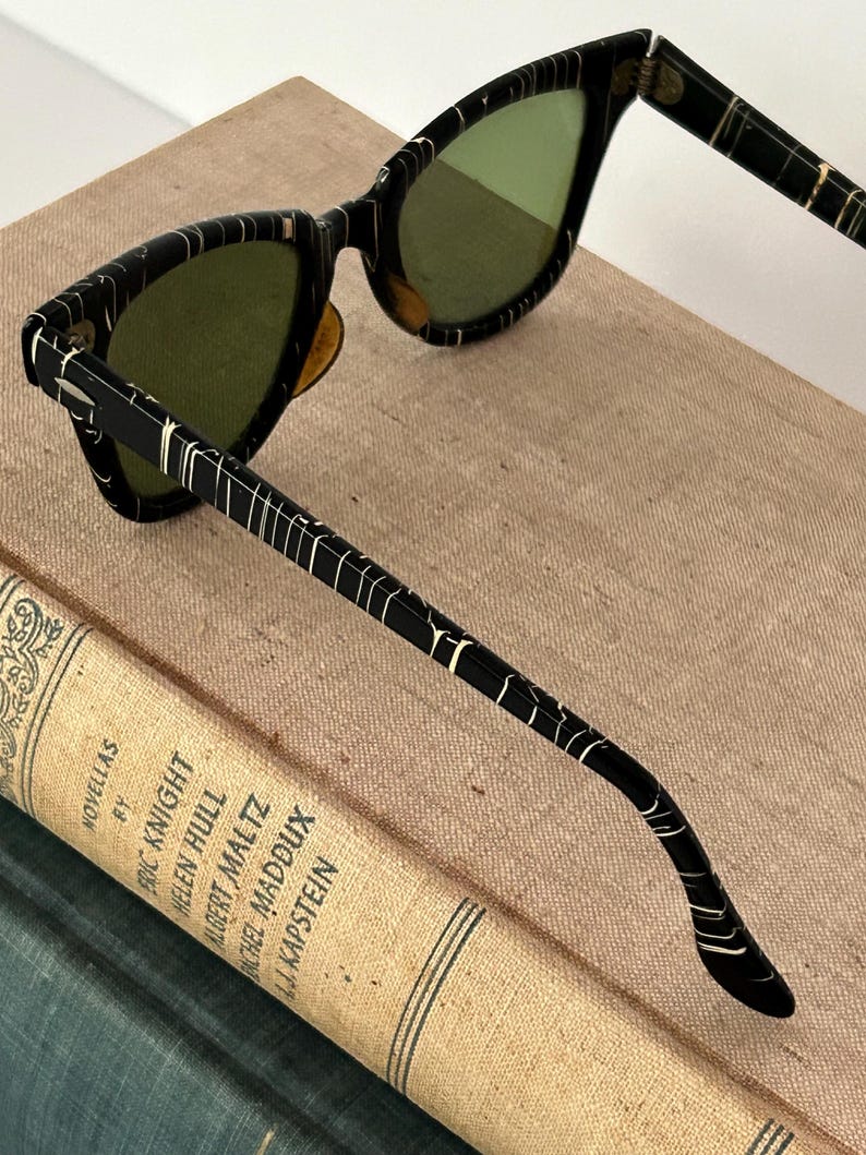 May include: Black cat-eye sunglasses with green lenses. The frames feature a unique pattern of white lines on a black background. The sunglasses are resting on a vintage book with the title "Novellas" and author names.