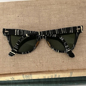 May include: Black and white patterned sunglasses with dark green lenses. The sunglasses have a retro design with a thick frame and a unique pattern. The glasses are resting on a tan surface.