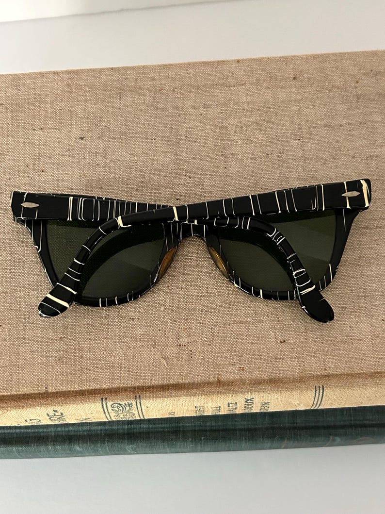 May include: Black and white patterned sunglasses with dark green lenses. The sunglasses have a retro design with a thick frame and a unique pattern. The glasses are resting on a tan surface.