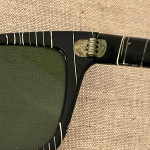 May include: Close-up of black sunglasses with a rectangular frame and green lenses. The frame has white vertical stripes along the top and sides. The hinge is gold-colored.