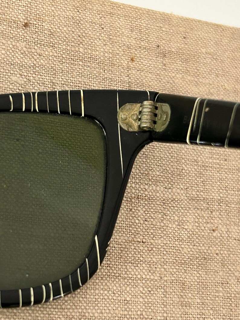 May include: Close-up of black sunglasses with a rectangular frame and green lenses. The frame has white vertical stripes along the top and sides. The hinge is gold-colored.