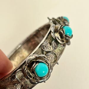 May include: A silver-toned hinged bangle bracelet featuring embossed floral and leaf patterns. The bracelet is decorated with three prominent, raised flower motifs, each centered with a smooth, round, bright turquoise-colored cabochon stone.
