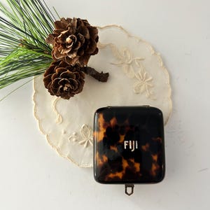May include: A tortoiseshell-patterned compact with the word "FIJI" in white lettering. The compact is square with rounded corners and a small metal clasp. The compact is on a white doily with floral embroidery, and next to it are two pine cones and pine needles.