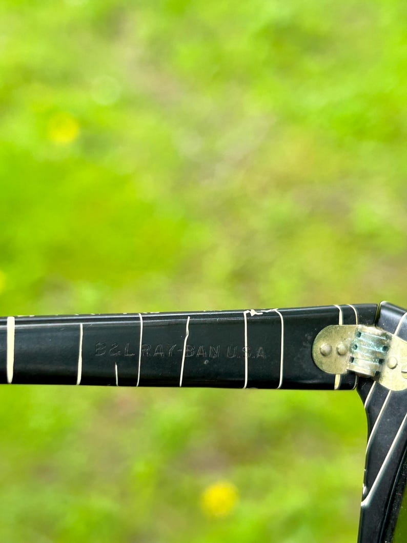 May include: Close-up of a black Ray-Ban USA sunglasses arm. The arm has white vertical lines and the text "B&L RAY BAN USA" in white. The hinge is gold-colored.