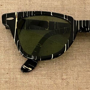 May include: Black sunglasses with a white striped pattern on the frames. The lenses are a dark green color. The sunglasses have a classic, retro design.