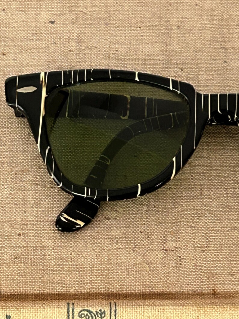 May include: Black sunglasses with a white striped pattern on the frames. The lenses are a dark green color. The sunglasses have a classic, retro design.