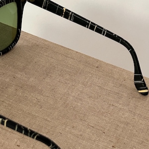 May include: Black sunglasses with a white striped pattern on the frames and arms. The lenses are a light green color. The sunglasses are resting on a tan surface.