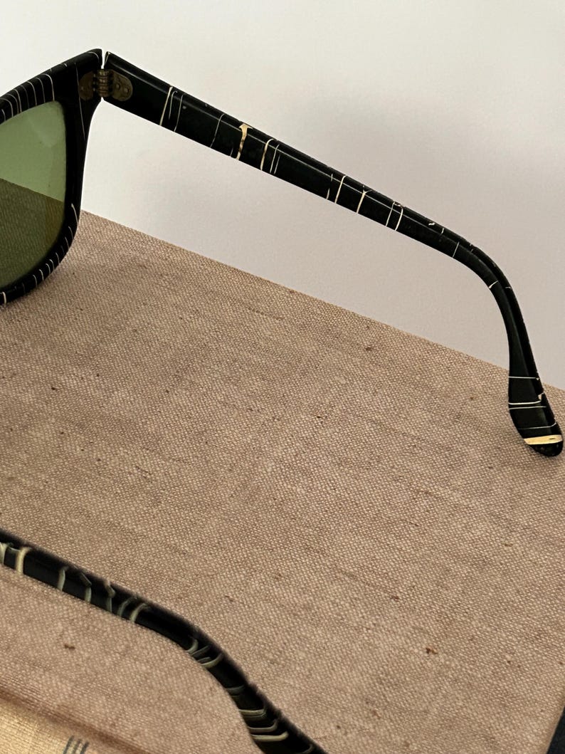 May include: Black sunglasses with a white striped pattern on the frames and arms. The lenses are a light green color. The sunglasses are resting on a tan surface.