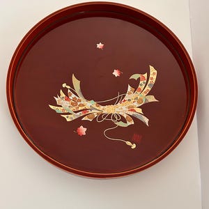 May include: A round, lacquered serving tray in a deep burgundy color. The tray features a decorative design of a stylized ribbon with gold, red, and green accents, along with small maple leaf motifs. A small, gold seal is visible on the right side.