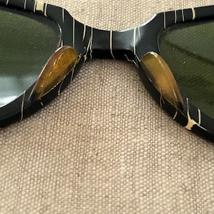 May include: Close-up of vintage sunglasses with a black frame and green lenses. The frame features a unique pattern of thin, gold-colored lines and accents. The lenses are a dark green color, providing a contrast to the black and gold frame.