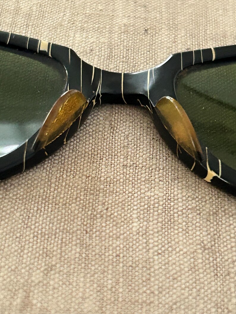 May include: Close-up of vintage sunglasses with a black frame and green lenses. The frame features a unique pattern of thin, gold-colored lines and accents. The lenses are a dark green color, providing a contrast to the black and gold frame.