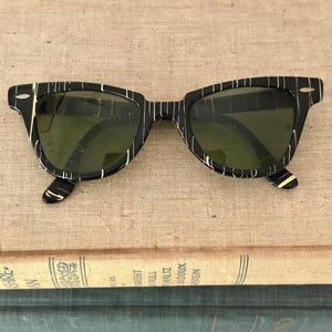 May include: Black sunglasses with a striped pattern in white and gold. The sunglasses have a classic square frame and green lenses. The glasses are resting on top of a stack of vintage books.
