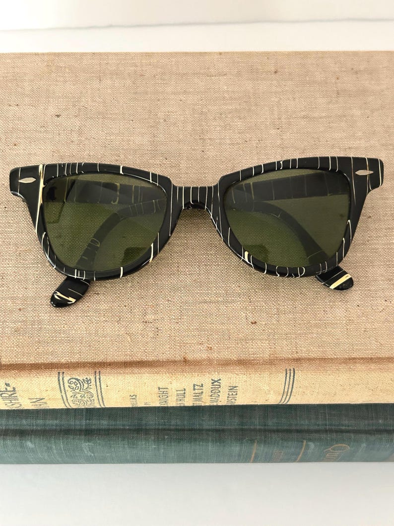 May include: Black sunglasses with a striped pattern in white and gold. The sunglasses have a classic square frame and green lenses. The glasses are resting on top of a stack of vintage books.