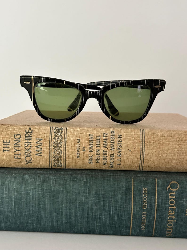 May include: Black and white striped sunglasses with green lenses rest on top of two vintage books. The sunglasses have a classic design with a thick frame and gold accents. The books are titled "The Flying Yorkshireman" and "Quotations."