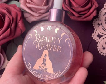 May include: A round, clear glass bottle with a pink metal cap and dropper. The bottle features a label with the text "Reality Weaver" and an illustration of a figure. The background includes pink and burgundy roses.