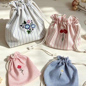 May include: Four fabric drawstring pouches featuring vertical stripes in blue, red, and pink. Each pouch is decorated with floral embroidery, including blue morning glories, red poppies, a pink carnation, and a small yellow daisy, all with white rope ties.