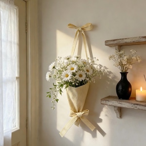 May include: A wall-hanging fabric cone in a yellow and white striped pattern, featuring a matching ribbon bow at the top and bottom. The cone holds a bouquet of white daisies and small white filler flowers, suspended against a plain wall.
