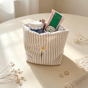 May include: A fabric storage basket featuring vertical beige and white stripes with a small embroidered white flower with a yellow center on the front. The basket is filled with various skincare products including a blue jar labeled Lancome.