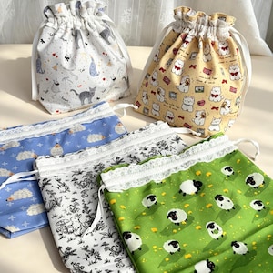 Peut inclure: Cinq pochettes en tissu à cordon de serrage avec divers motifs : un sac blanc avec des chats gris, un sac jaune avec des chiens blancs, un sac bleu avec des hérissons, un sac blanc avec des illustrations de forêt noire et un sac vert avec des moutons, tous avec une bordure en dentelle blanche.