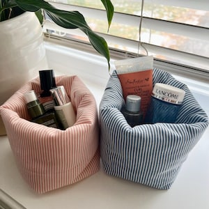 May include: Two fabric storage baskets with a soft, quilted texture. One basket features a pink and white pinstripe pattern, while the other has a blue and white pinstripe pattern. Both contain various skincare bottles and tubes.