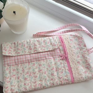 May include: A floral print clutch with a pink gingham flap and strap. The clutch features a pink zipper, lace trim, and a small bow. A clear glass candle sits in the background.