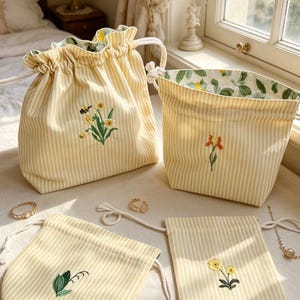Puede incluir: Una colección de bolsas y bolsitas con cordón a rayas amarillas y blancas. Cada bolsa presenta diseños florales bordados, con un forro floral verde. Varios anillos y joyas de oro están esparcidos alrededor de las bolsas.