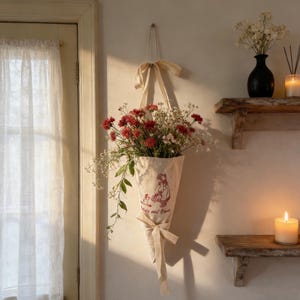 May include: A cream canvas flower holder with a red illustration and text, suspended on a wall. It's filled with red and white flowers and greenery. Cream ribbons are tied at the top and bottom, creating a decorative display.