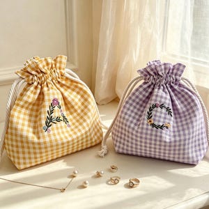 May include: Two drawstring pouches made of gingham fabric, one in yellow and one in lavender, each featuring a floral embroidered letter. The yellow pouch displays the letter A, while the lavender pouch displays the letter C. White rope cords close the bags.