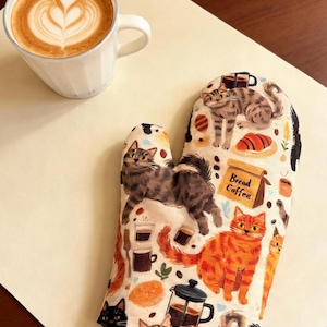 May include: A white ceramic mug with a latte and heart-shaped foam art sits beside a patterned oven glove. The oven glove features a cat and coffee theme with the words "Bread Coffee".
